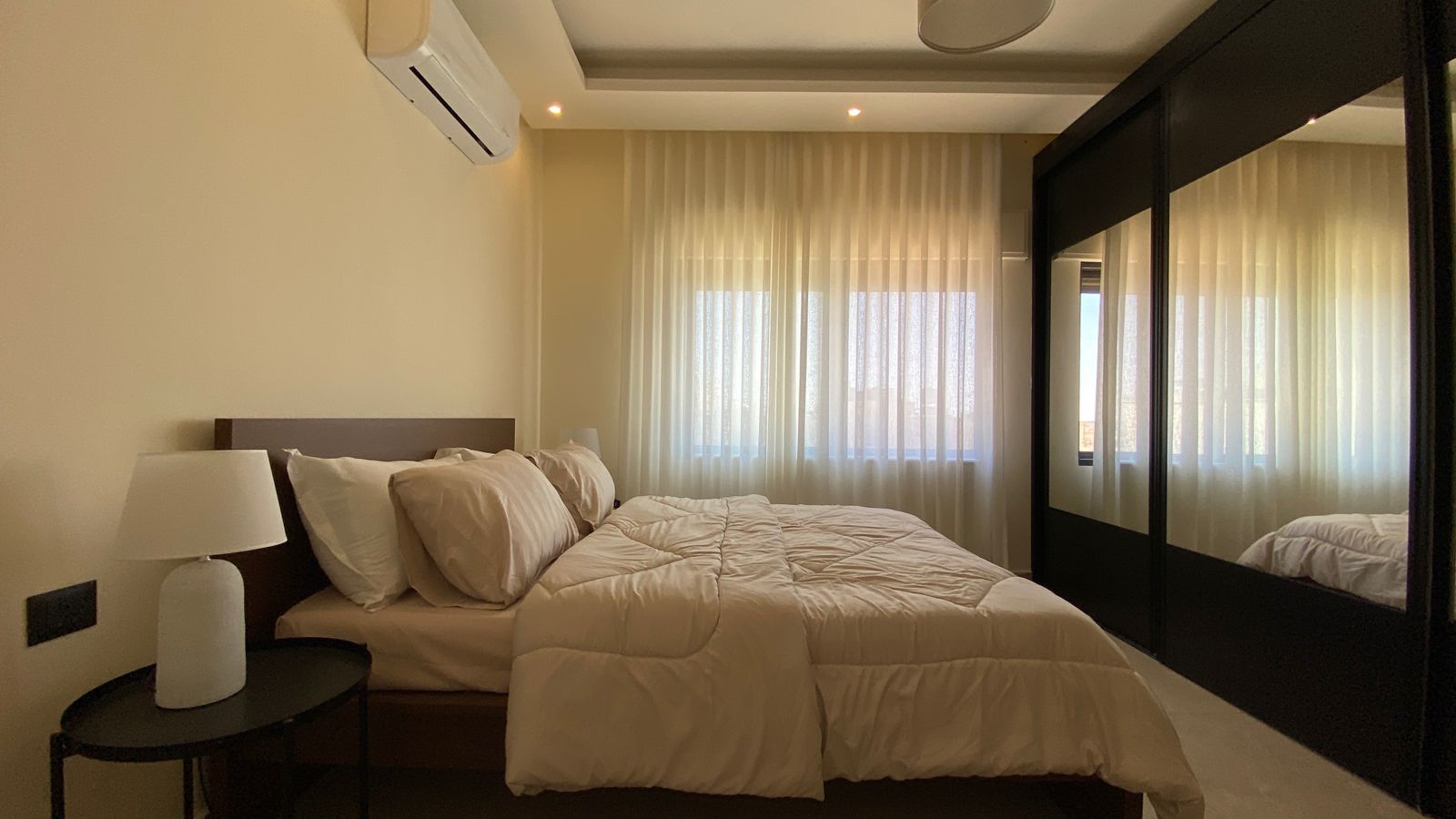 56bb6868-008c-4b5d-9c59-1d1be6e2b640 Modern hotel-style bedroom with a white quilted bed, two pillows, and a dark wood headboard beside a round black nightstand and white lamp; sheer curtains over a large window across the room.