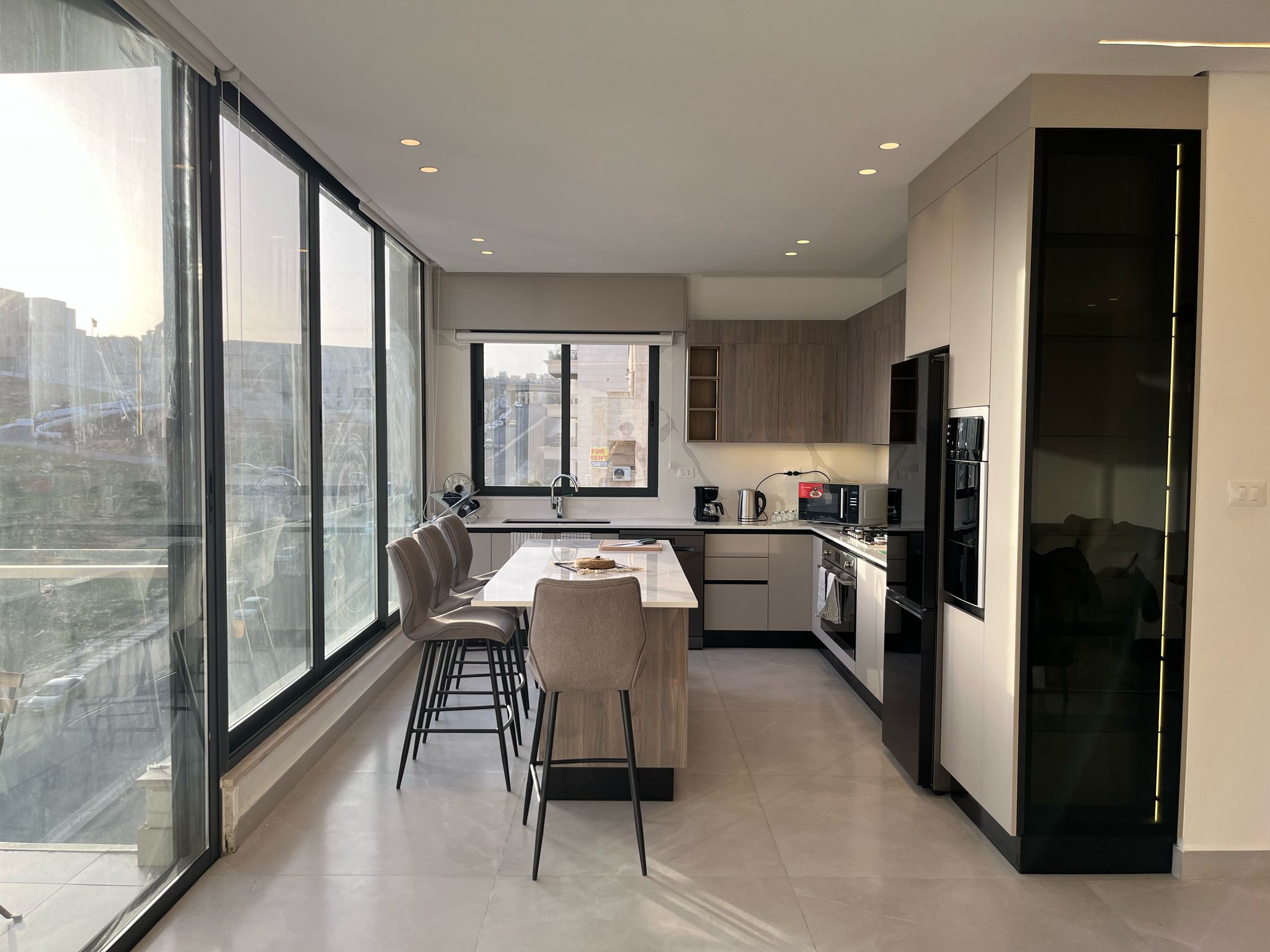 57741c6a-e9c8-4609-b735-982f7dcc1566 Bright, modern kitchen with a long white island, beige bar stools, and large glass windows along the wall letting in daylight.