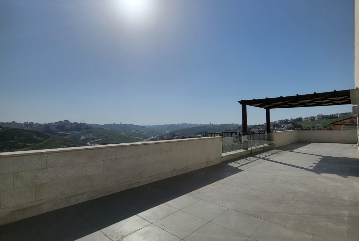 Sunny rooftop terrace with large gray tiles, concrete railing, and a distant hillside town under a clear blue sky.