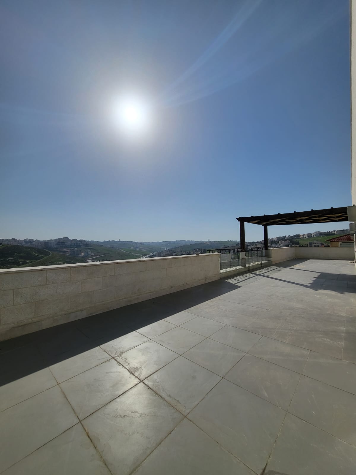 581739dd-38ac-484c-80e6-b242c3ef53c8 Sunny rooftop terrace with large gray tiles, concrete railing, and a distant hillside town under a clear blue sky.