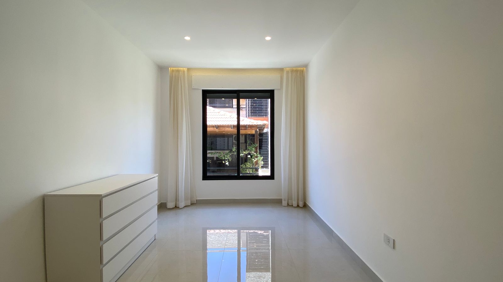 58ecfa5e-1063-4e61-be60-e58d8ca24392 Bright minimalist room with white walls and glossy floor; a white dresser sits on the left, a large window with sheer curtains at the far end.