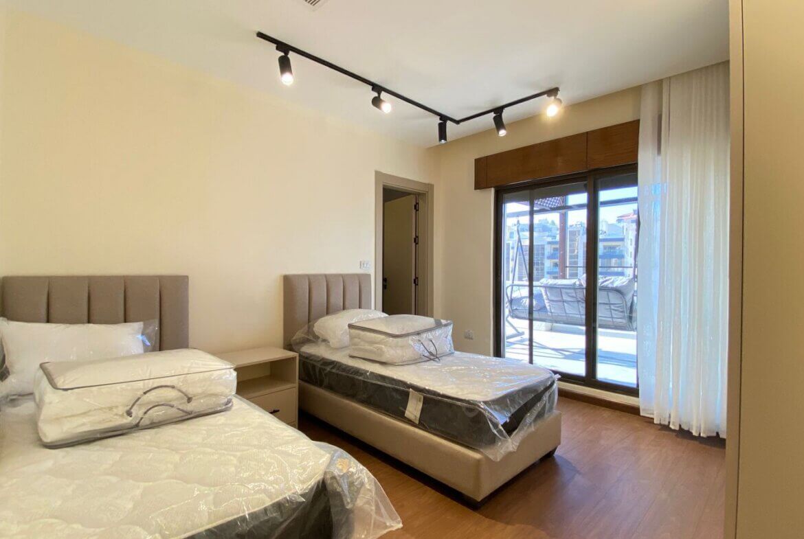Two beige upholstered twin beds with plastic-wrapped mattresses, a shared nightstand between them, in a bright bedroom with wooden floor.