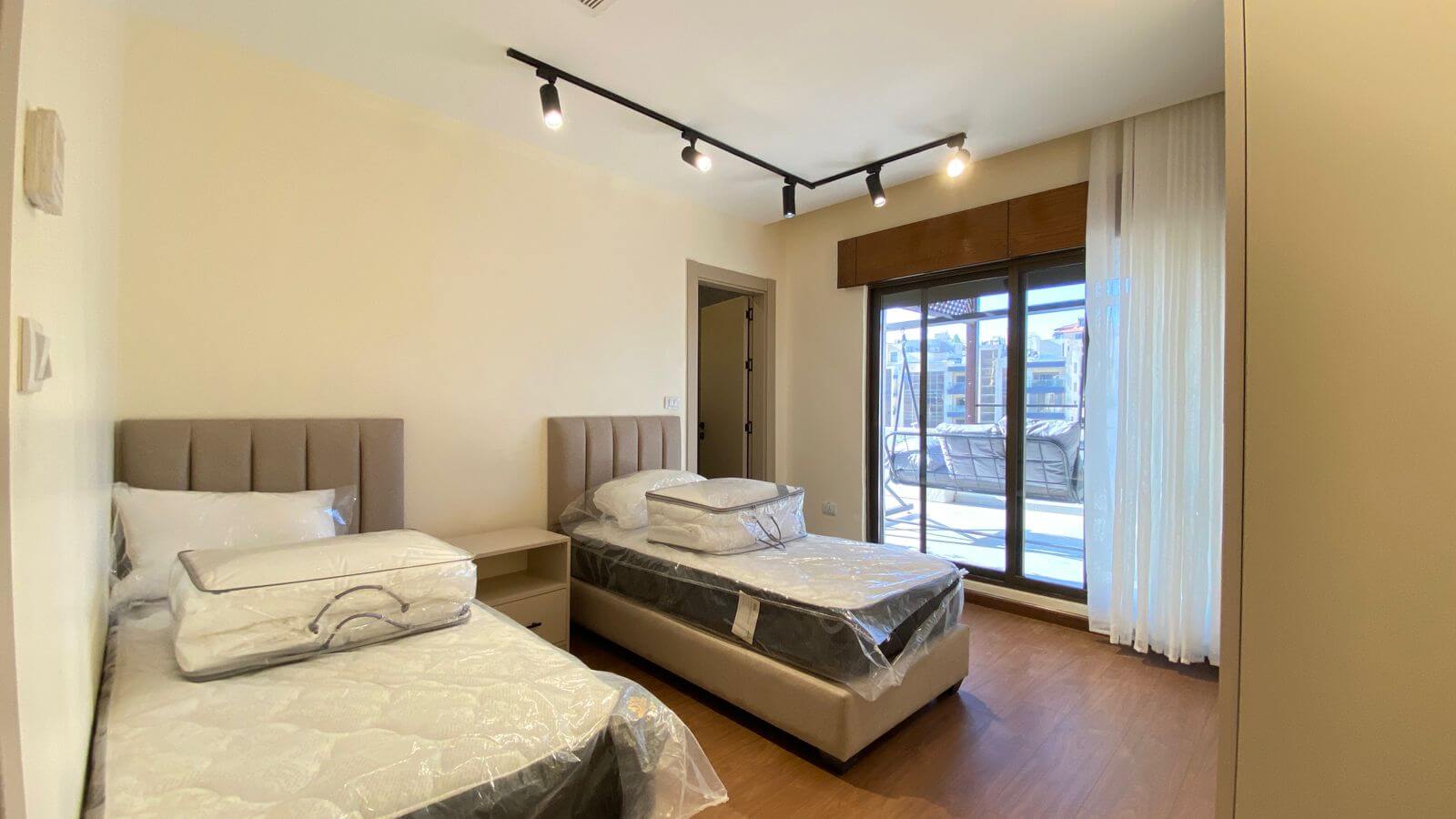 Two beige upholstered twin beds with plastic-wrapped mattresses, a shared nightstand between them, in a bright bedroom with wooden floor.