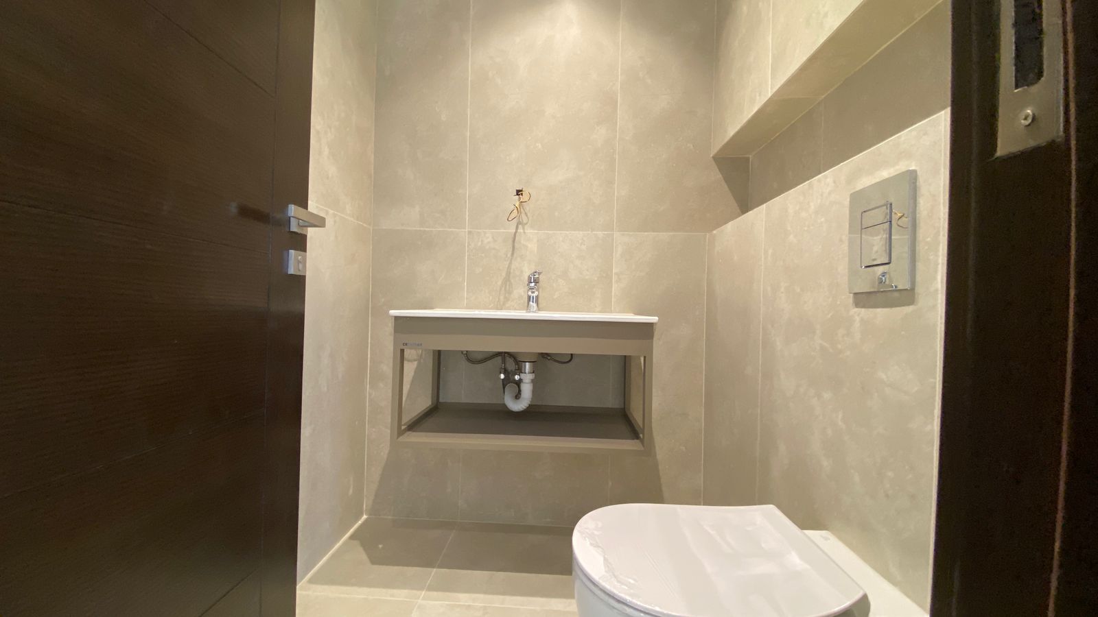 5957ea59-3d6c-4970-a92f-7afef63958c6 Compact bathroom with beige tile walls, a wall-mounted sink over exposed plumbing, and a white toilet in the foreground.