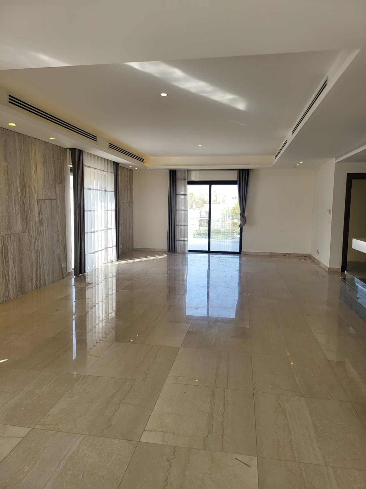 595f20b9-a0cc-443e-b482-d40f6addf813 Spacious modern living room with glossy beige tile floor, marble paneled wall on left, and large sliding glass doors to a balcony letting in daylight.
