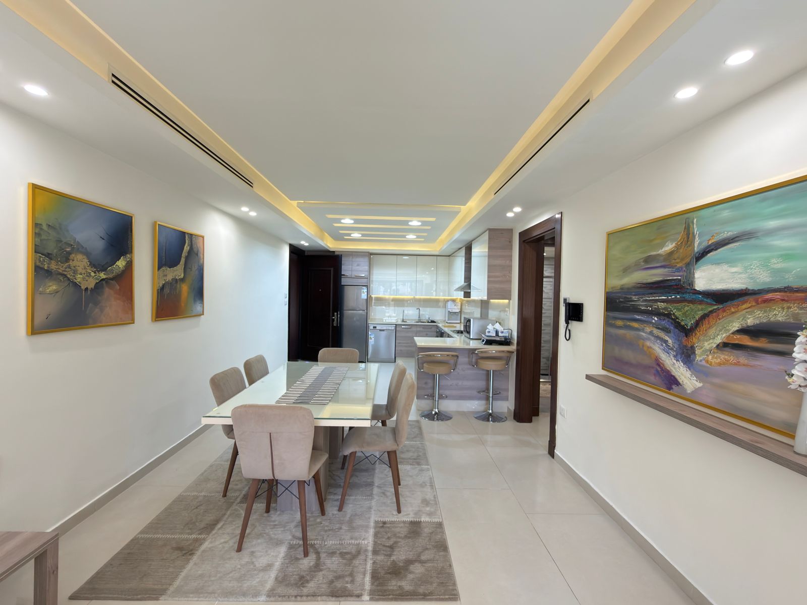 5a242349-bbd5-46d2-bf71-6bc58150157d Open-concept dining area with a glass-top table, six beige upholstered chairs, a gray rug, and a modern kitchen in the background. Artistic wall pieces line the left wall while a large colorful painting adorns the right wall.