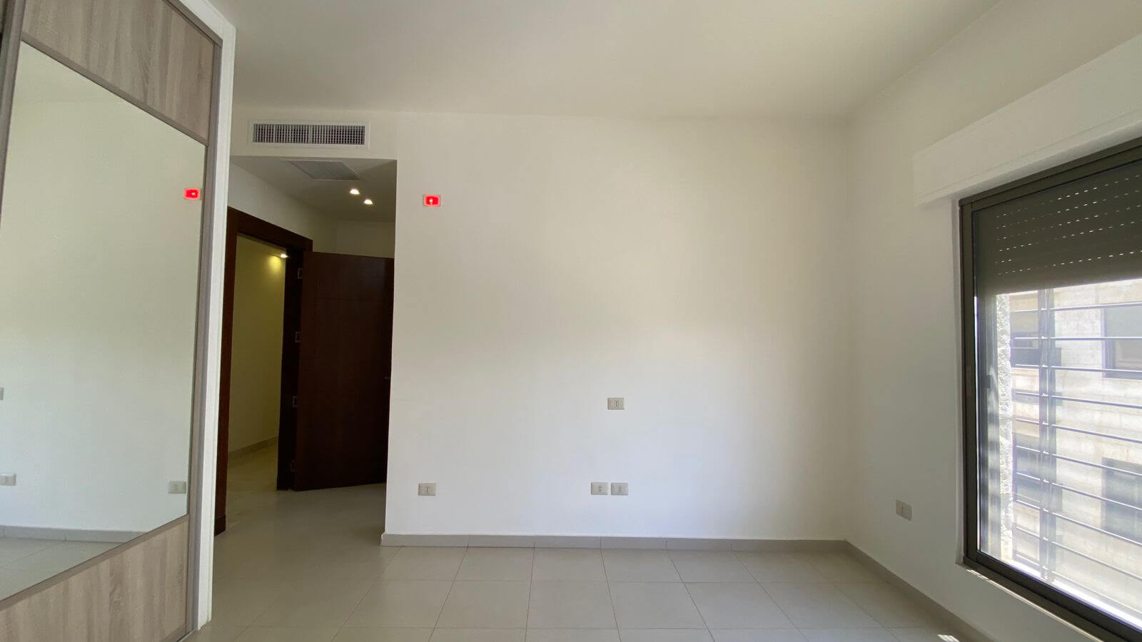 5a6dfaa0-33f1-4506-8b03-6ace1742e0fc Empty, modern room with a large window on the right, tiled floor, and a mirrored built-in wardrobe on the left.