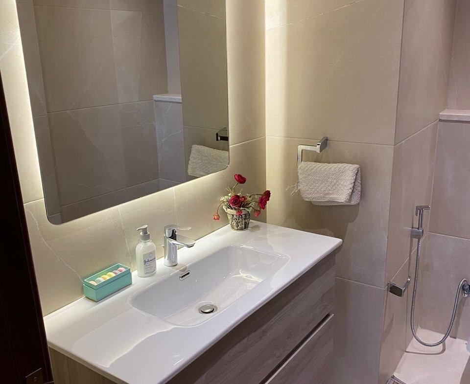 Modern bathroom vanity with a white sink, wood cabinet, backlit mirror, soap dispenser, turquoise toiletries tray, and a small vase of red flowers.
