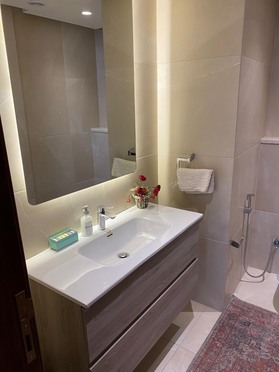 5b005c9c-de57-4d75-b93d-e0725cfab027 Modern bathroom vanity with a white sink, wood cabinet, backlit mirror, soap dispenser, turquoise toiletries tray, and a small vase of red flowers.