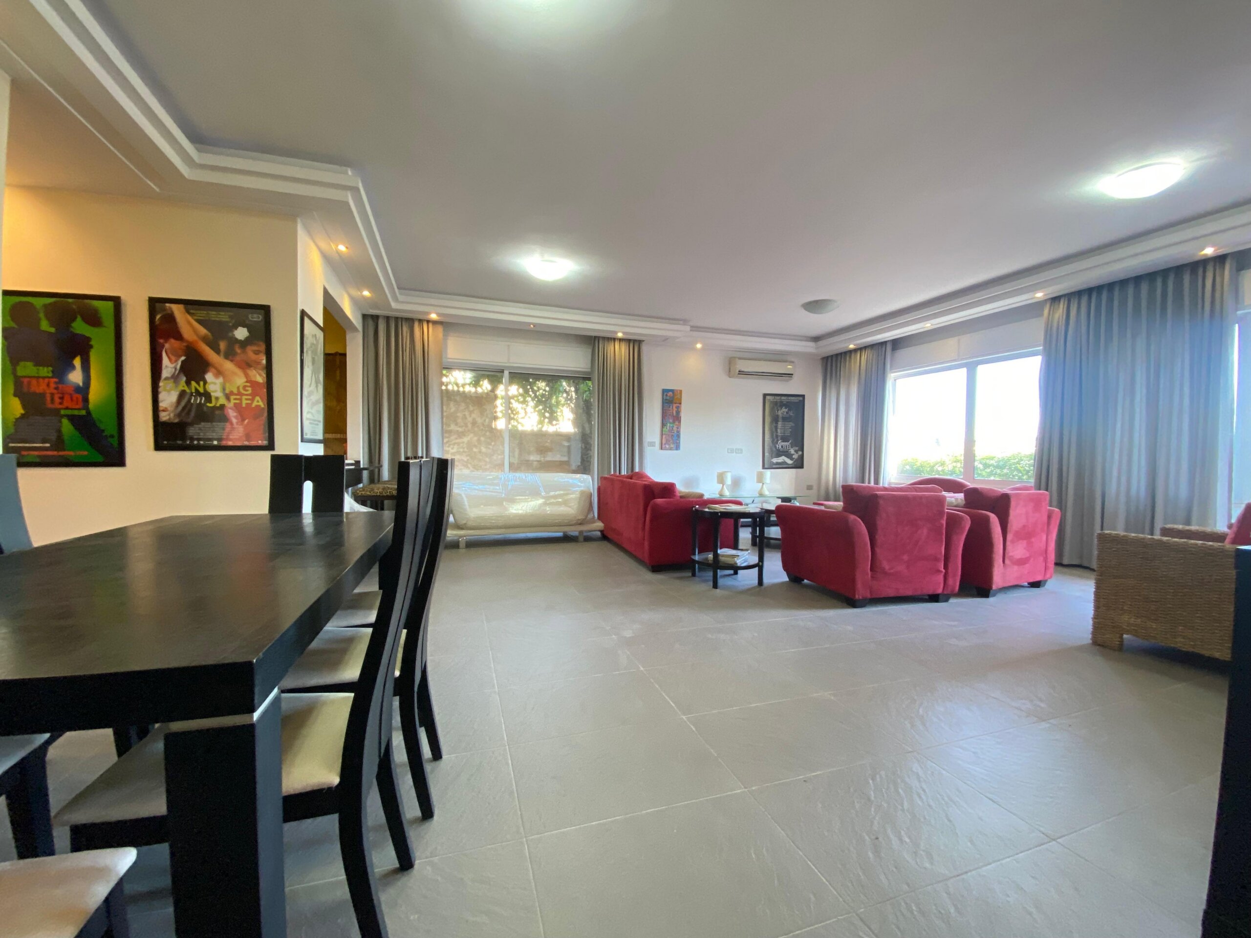 5b34b632-536c-4c56-9a51-cdc643f82682 Spacious living room with black dining table and chairs on the left, red couches in a bright lounge with posters on the wall and large windows with curtains on the right