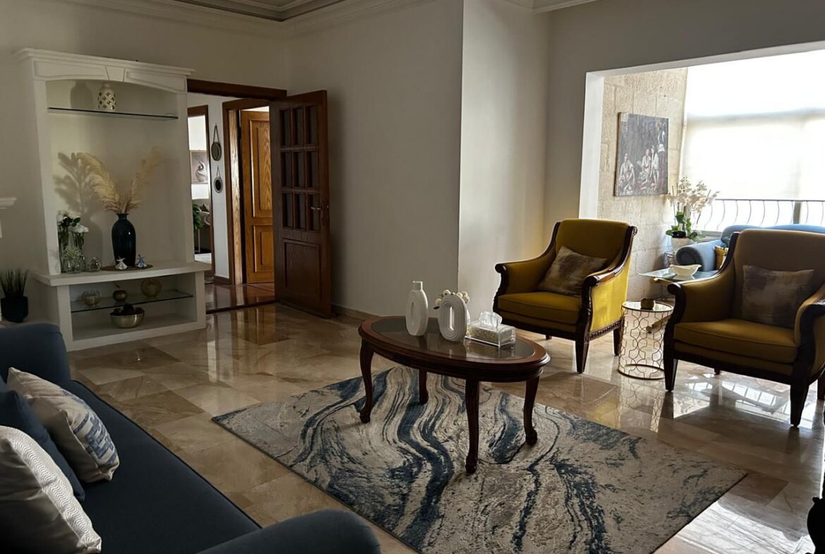 Bright living room with a patterned area rug, a wooden coffee table, and yellow armchairs by a balcony window; blue sofa on the left.”