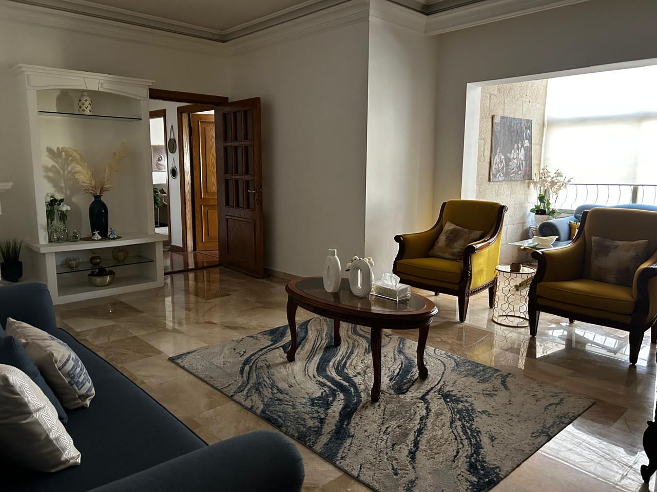 5b70e062-30b2-45a5-8b43-29ebb3da28a5 Bright living room with a patterned area rug, a wooden coffee table, and yellow armchairs by a balcony window; blue sofa on the left.”