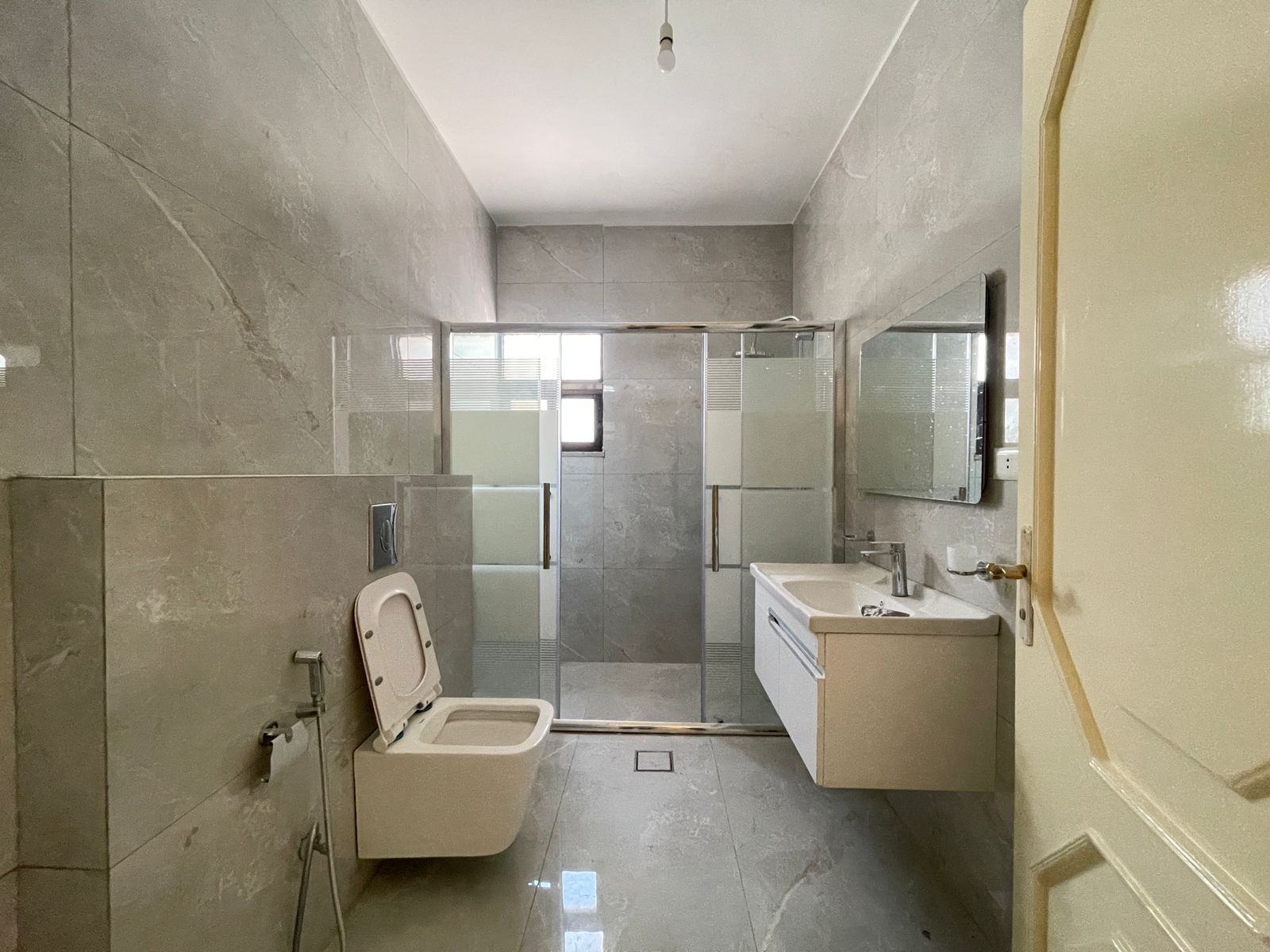 5b8430b5-060c-475d-b510-d07aff2bdfa0 Grey-tiled bathroom with a wall-hung toilet on the left, a glass shower enclosure in the back, and a white vanity with mirror on the right.