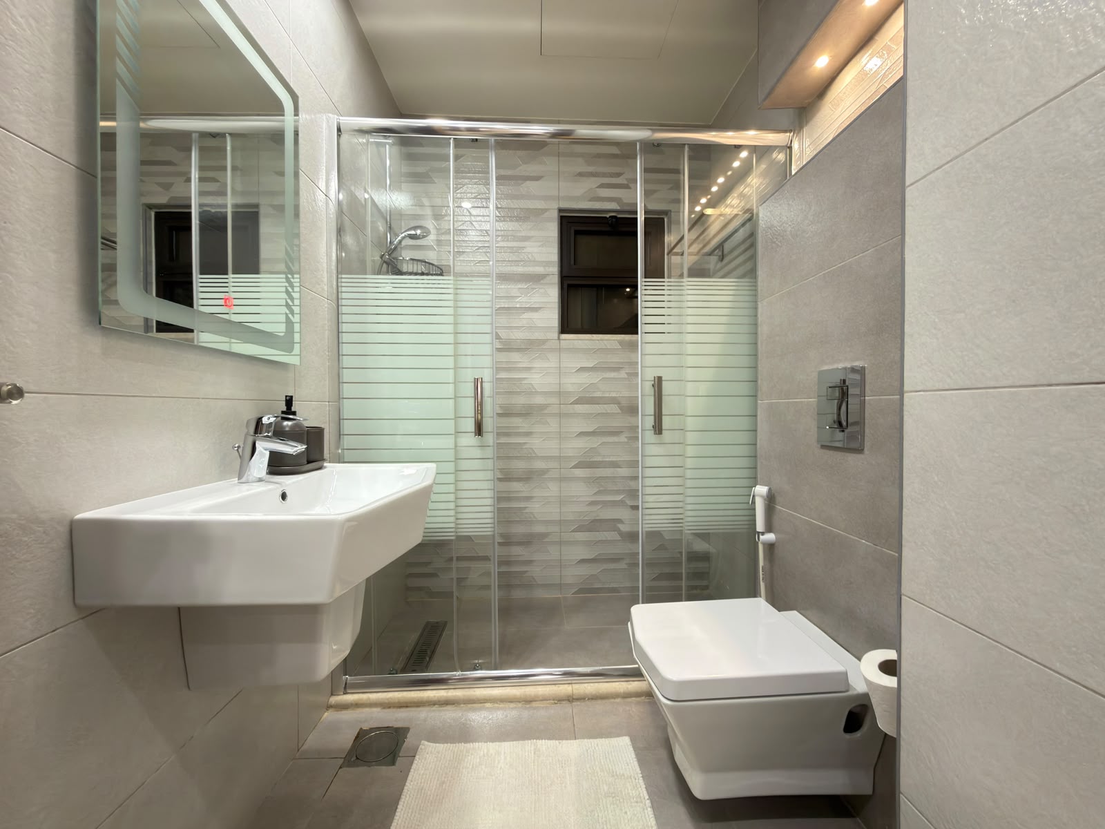 5d12451c-1d39-493a-8cad-8a982eb96a99 Modern bathroom with a glass-framed shower enclosure, wall-mounted sink, and wall-hung toilet in a sleek gray tile setting.
