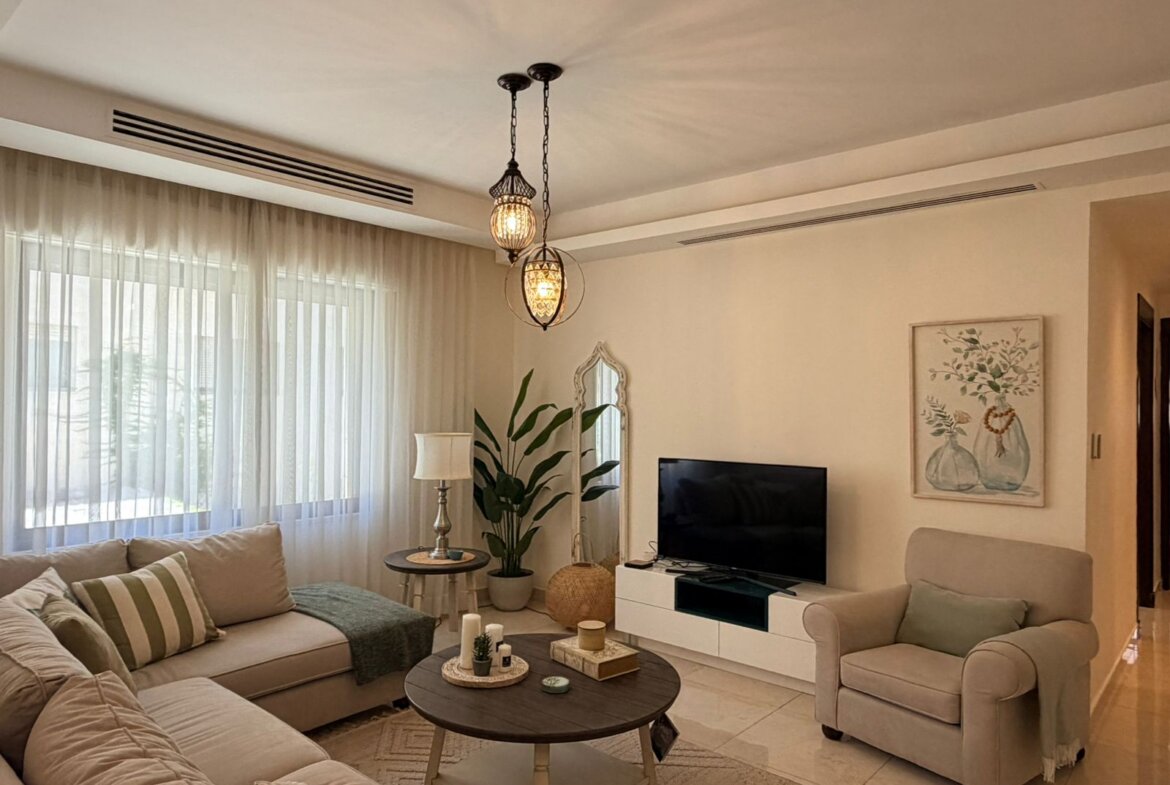Neutral-toned living room with beige sofa and armchair, round coffee tables, and large windows with sheer white curtains letting in daylight, plus a decorative chandelier overhead.