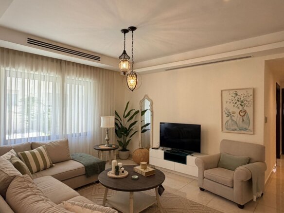 Neutral-toned living room with beige sofa and armchair, round coffee tables, and large windows with sheer white curtains letting in daylight, plus a decorative chandelier overhead.