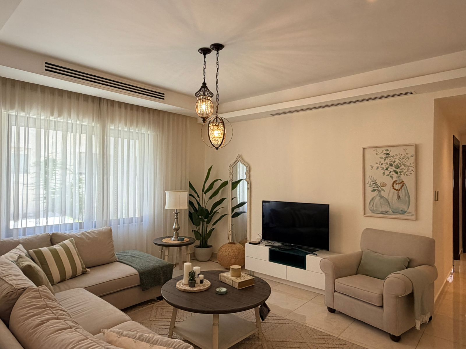 5d42490c-21da-4eb1-b90e-790053a7566b Neutral-toned living room with beige sofa and armchair, round coffee tables, and large windows with sheer white curtains letting in daylight, plus a decorative chandelier overhead.