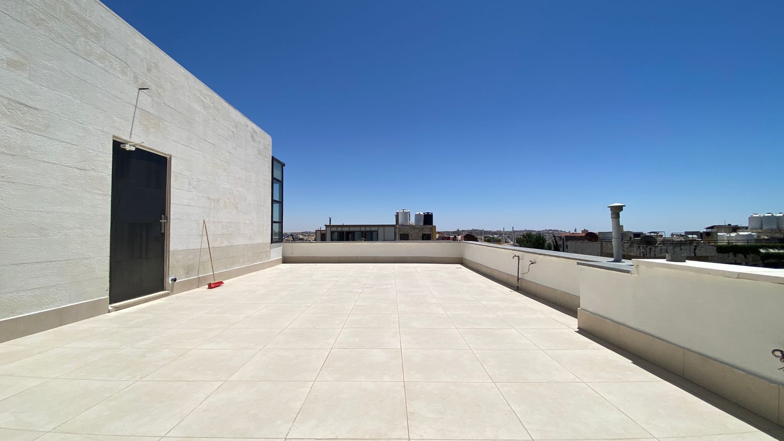 5d574934-43c0-4464-a2e1-41b555bfad48 Sunny rooftop terrace with pale walls and a left-side door; a broom resting by the door, and a distant city skyline under a blue sky.