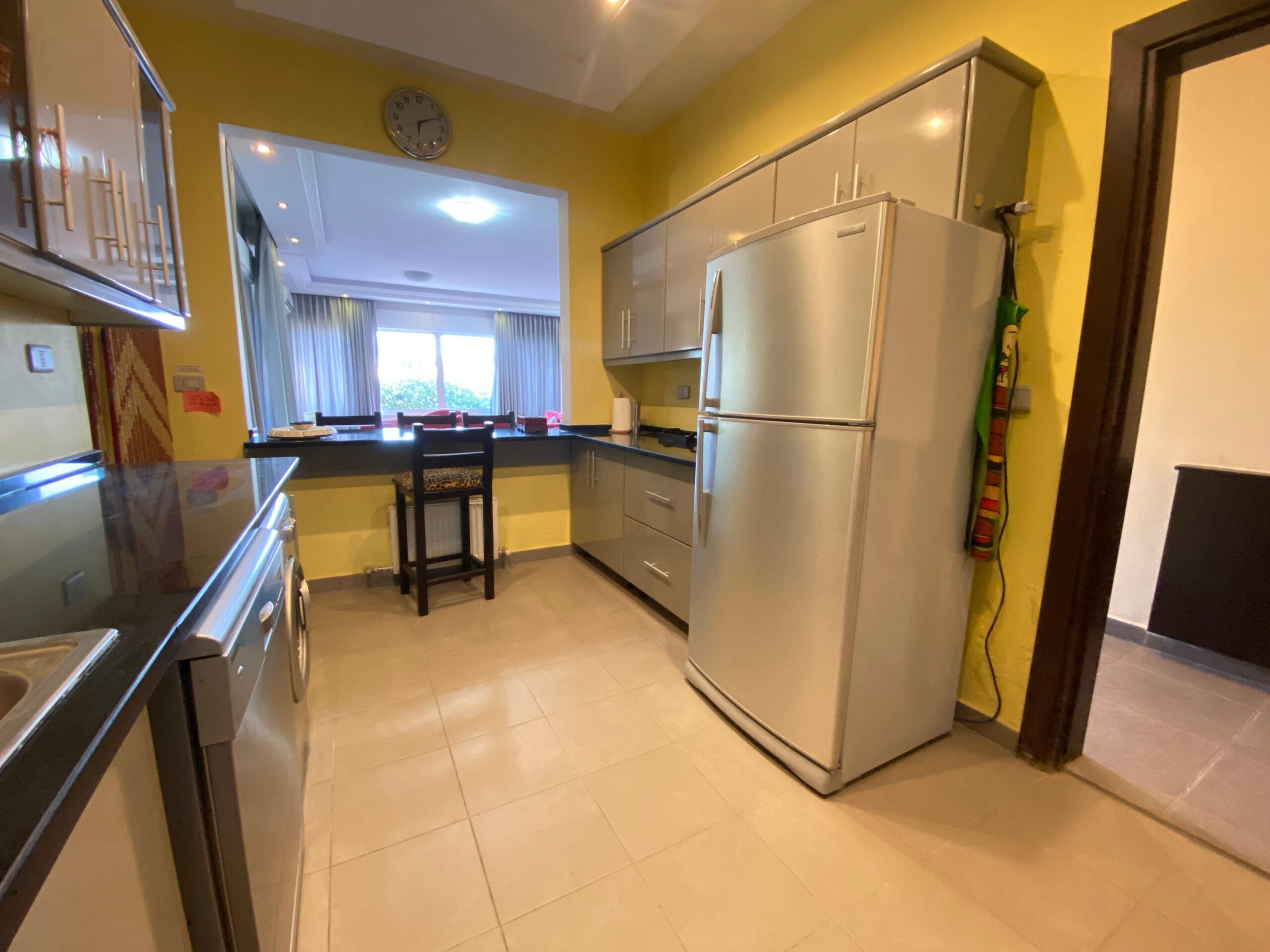5d8cc84f-f416-47eb-b7c6-a90b28c2ba55 Modern kitchen with stainless steel fridge and yellow walls; dining area visible through doorway with chairs and windows in background.