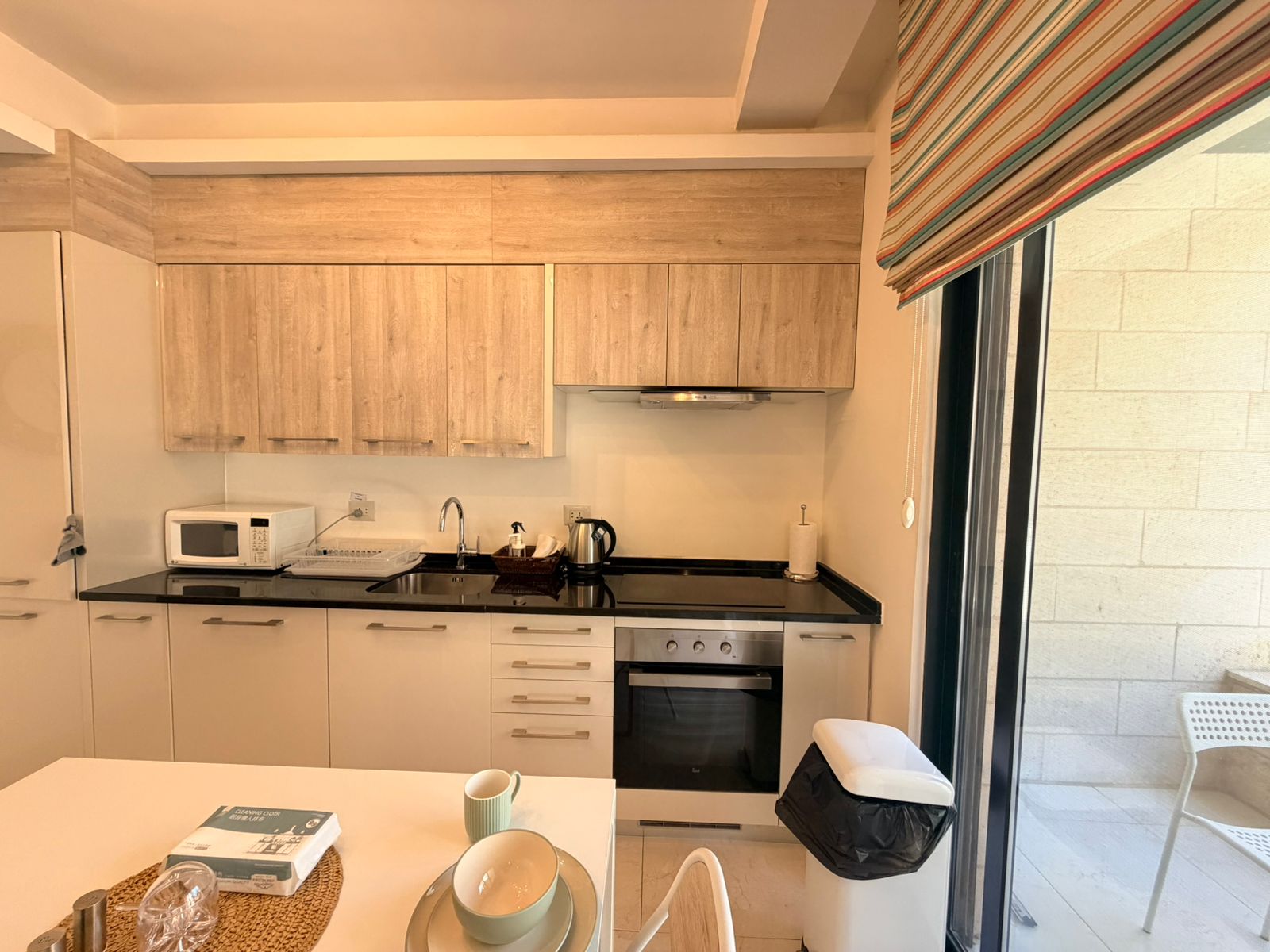 5e7ceb56-2cba-40e4-8d19-a14d7ca59899 Modern kitchen with light wood cabinets, black countertop, and stainless steel oven; kettle and microwave on counter.