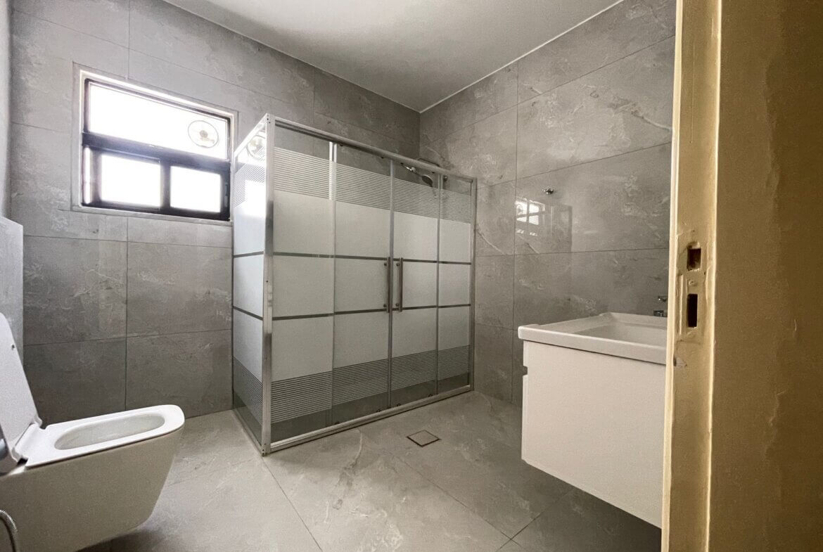 5f7987fb-e654-42f3-8e9d-9e8c4b7e8a32 Gray marble-tiled bathroom with a frosted glass shower enclosure, a white toilet, and a white vanity under a small window on the left side.