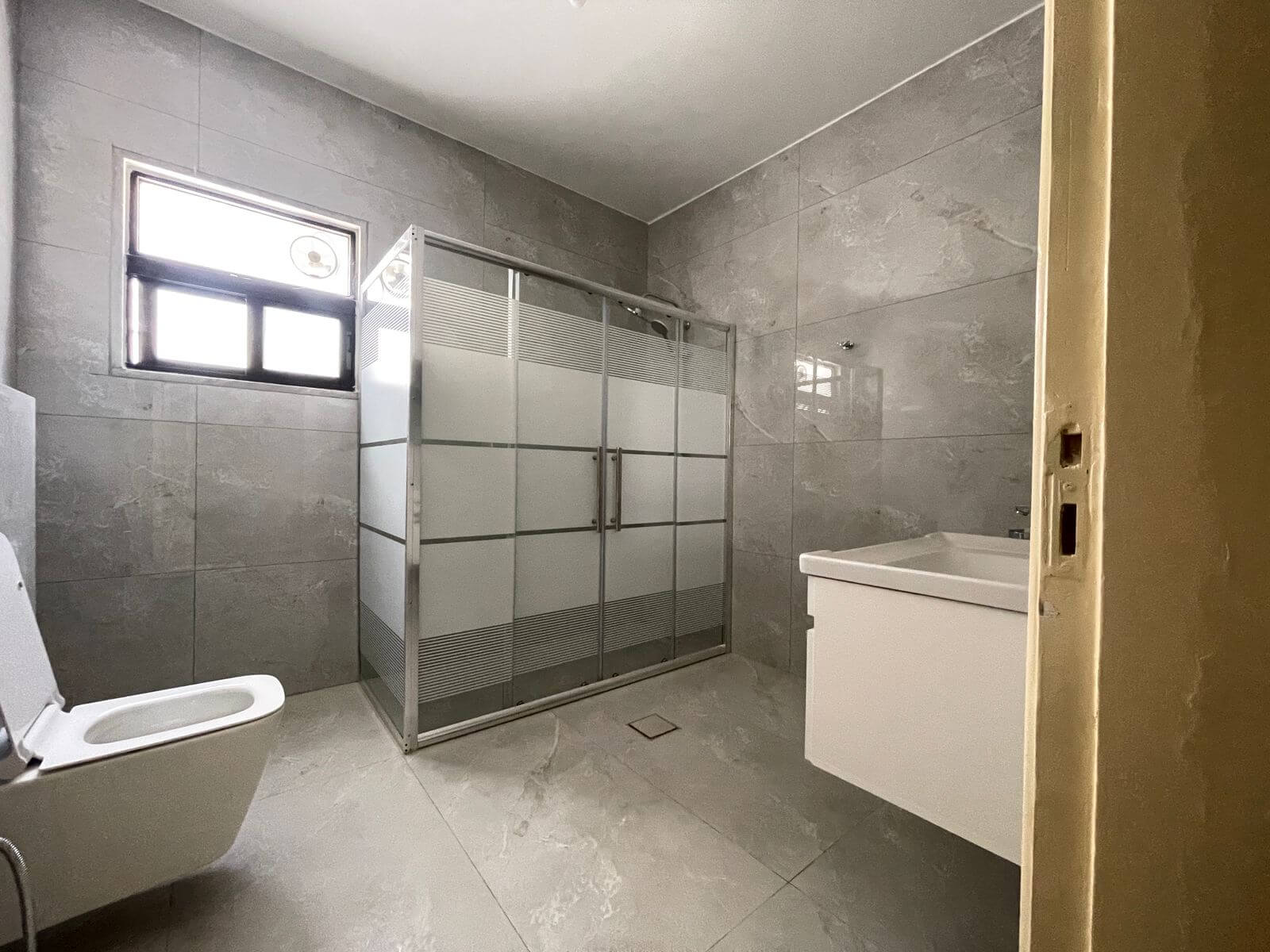 5f7987fb-e654-42f3-8e9d-9e8c4b7e8a32 Gray marble-tiled bathroom with a frosted glass shower enclosure, a white toilet, and a white vanity under a small window on the left side.