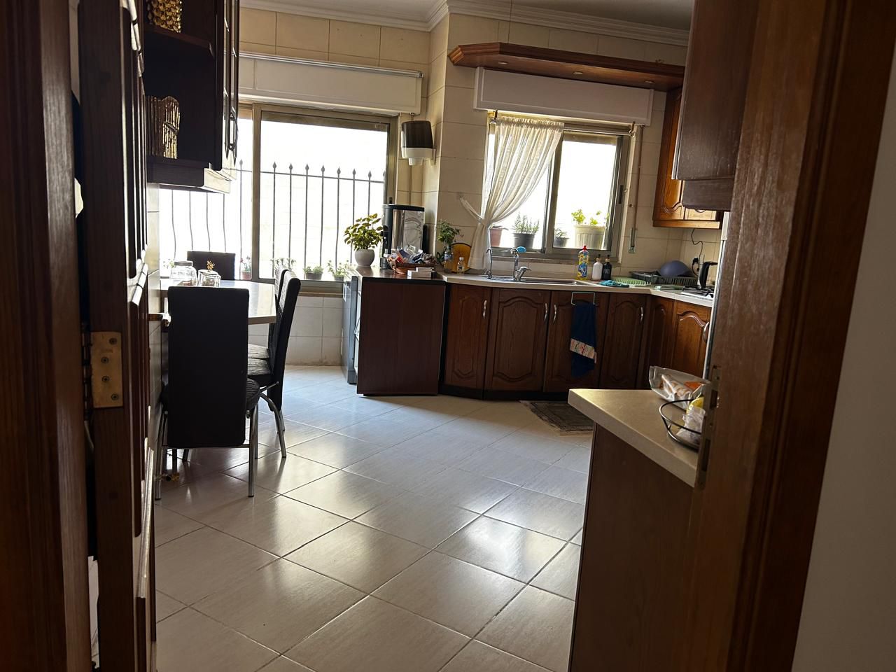 6088228e-54b5-4ab3-90bb-135dcd1d9f93 Sunlit kitchen with dark wooden cabinets, a dining table and chairs by a window with potted plants on the sill.