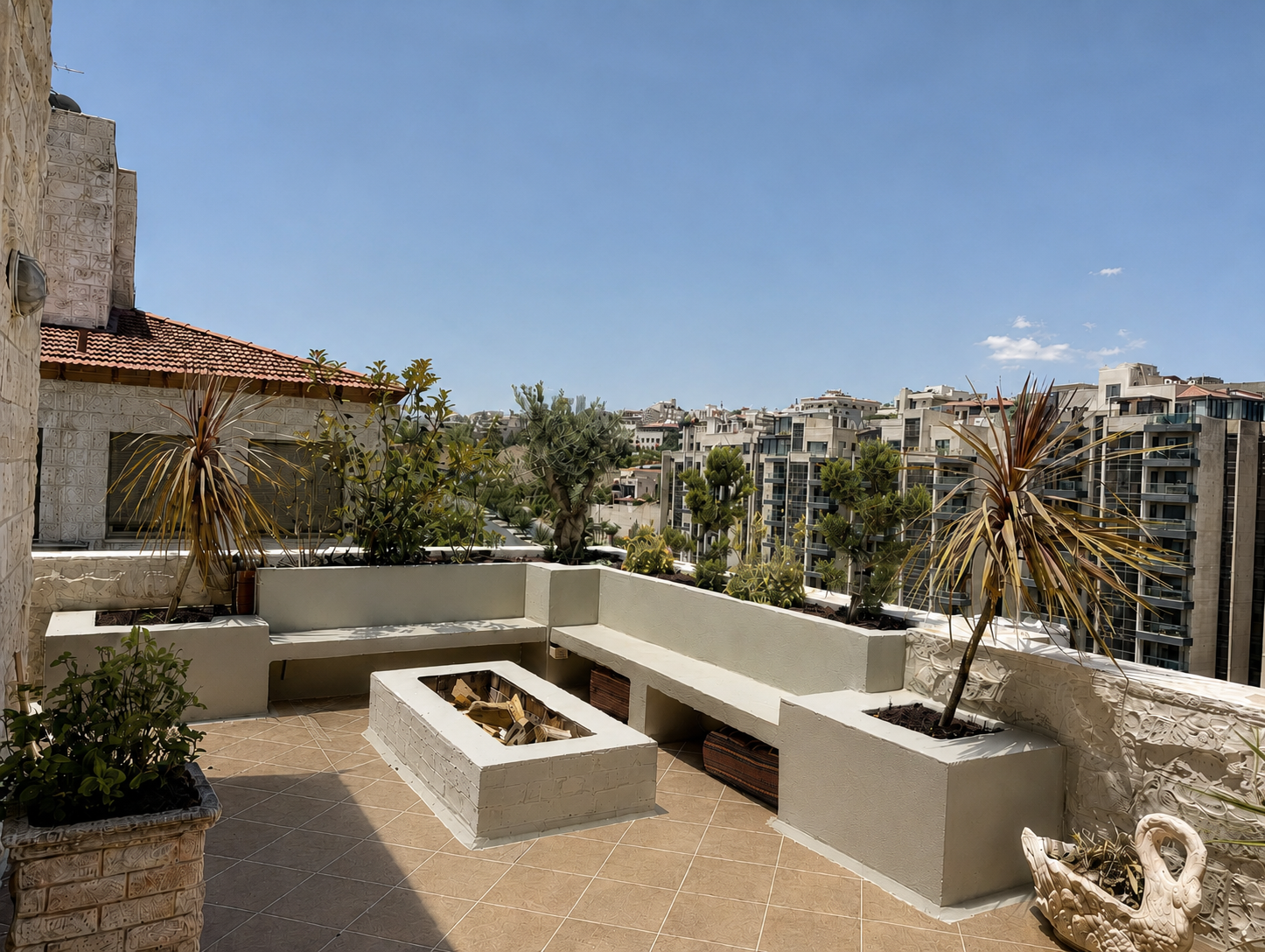 6149d31d-6512-49e7-80ac-f343fcbb2221 Rooftop terrace with built-in benches and raised planters, green plants, and a cityscape of modern apartment buildings under a clear blue sky.