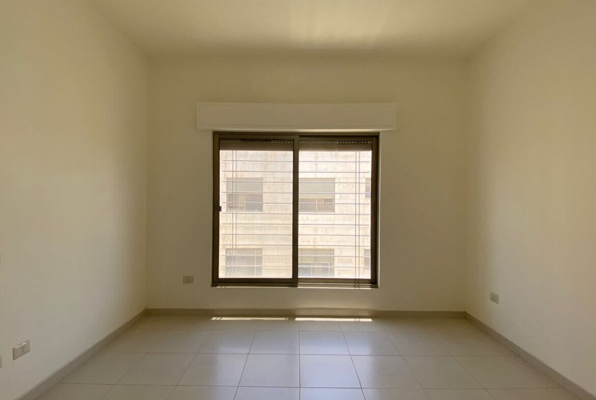 62290d43-5101-44e2-a365-a8d21885ec6a Empty, neutral-toned room with a large central window; sunlight pools on the tiled floor, view of a building outside.