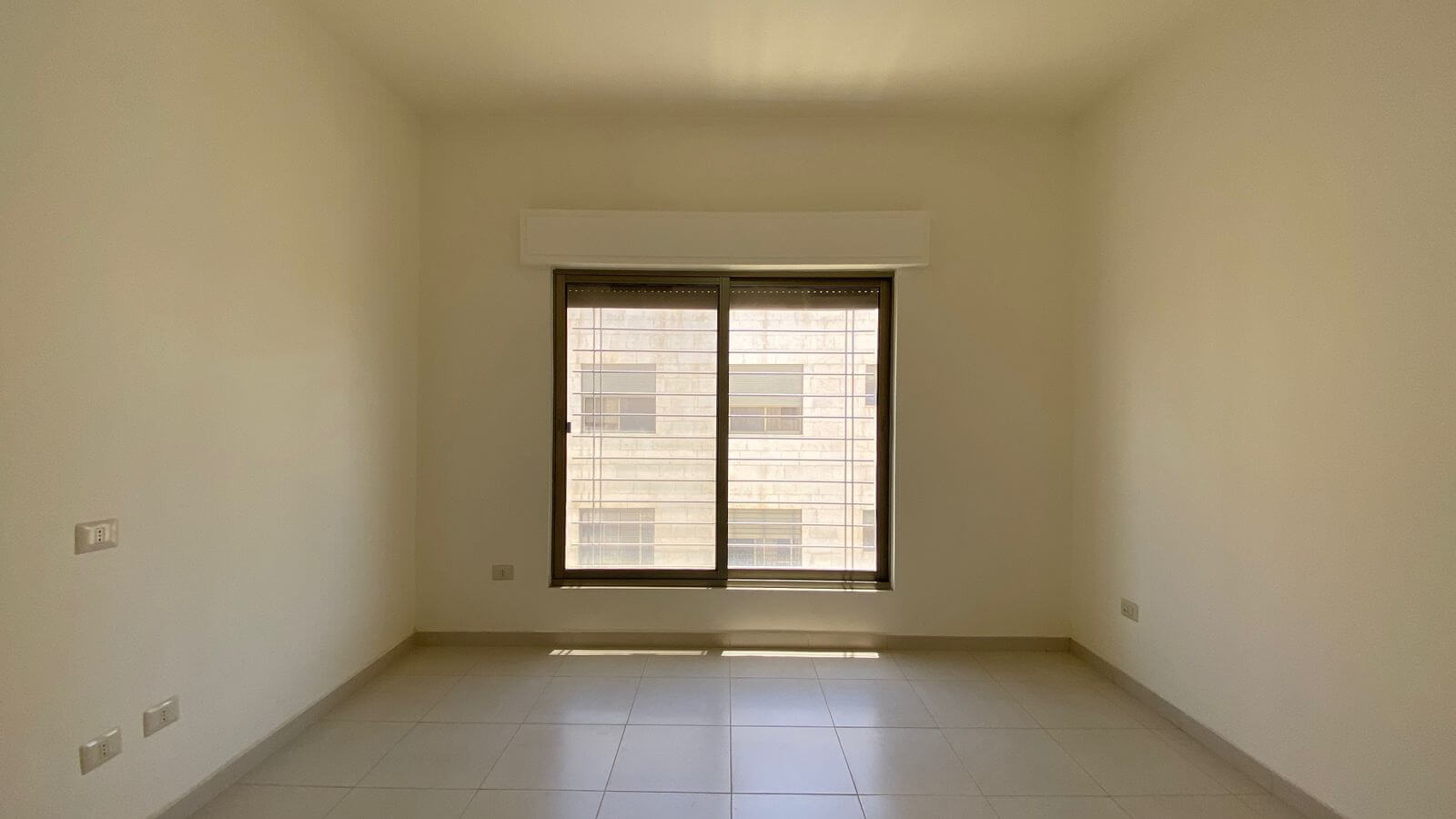 62290d43-5101-44e2-a365-a8d21885ec6a Empty, neutral-toned room with a large central window; sunlight pools on the tiled floor, view of a building outside.