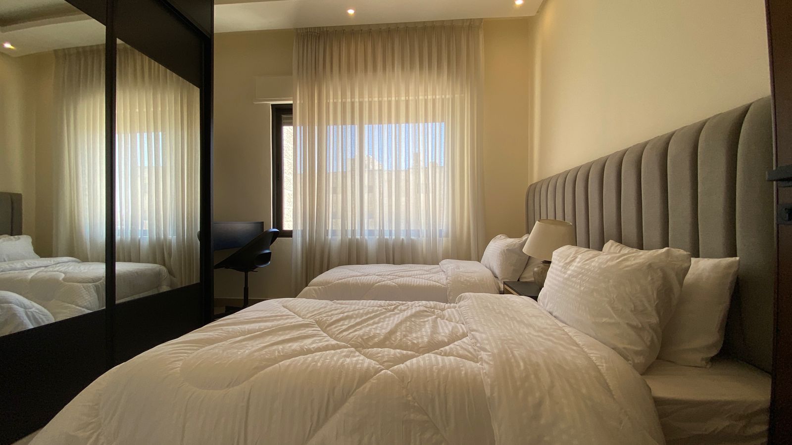 62d562df-e944-4526-a114-3f3aec778aa7 Hotel room with two twin beds, white quilts, and a tufted gray headboard by a window with sheer curtains.