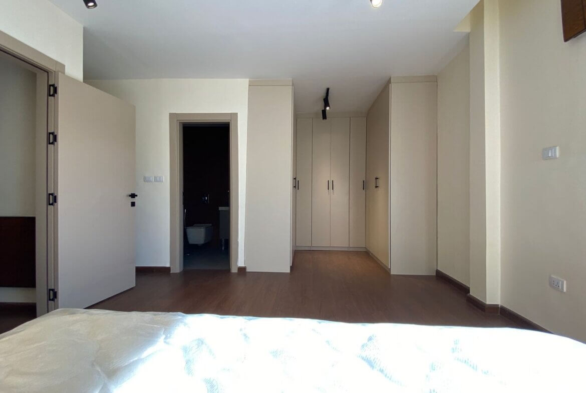 Modern bedroom with built-in beige wardrobes along the back wall, a doorway to a dark bathroom on the left, and a bed in the foreground.