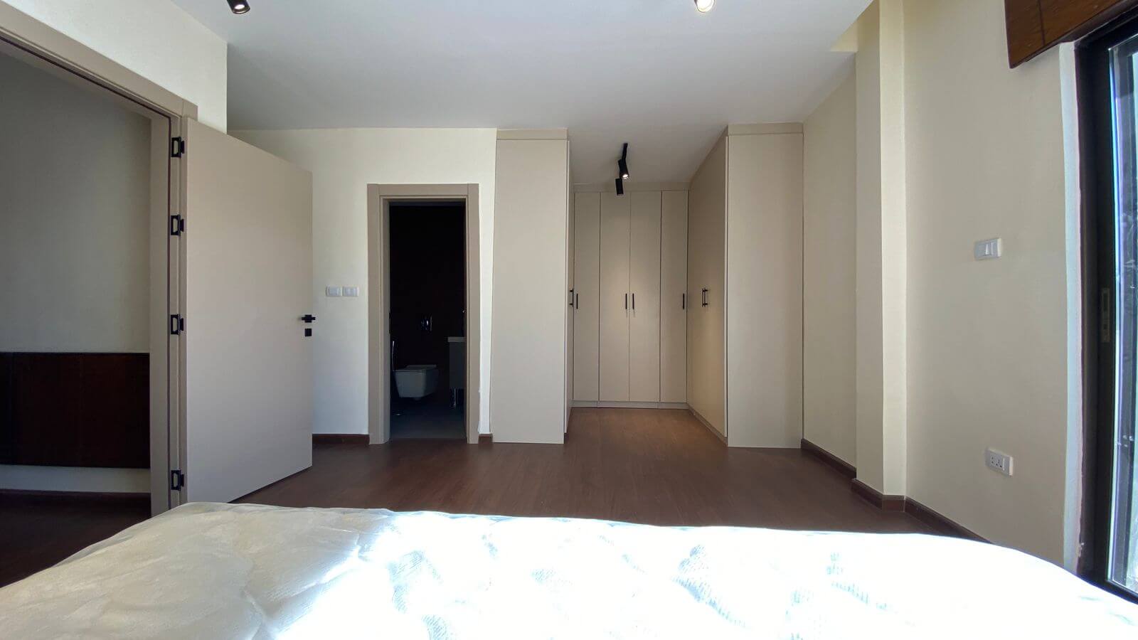 Modern bedroom with built-in beige wardrobes along the back wall, a doorway to a dark bathroom on the left, and a bed in the foreground.