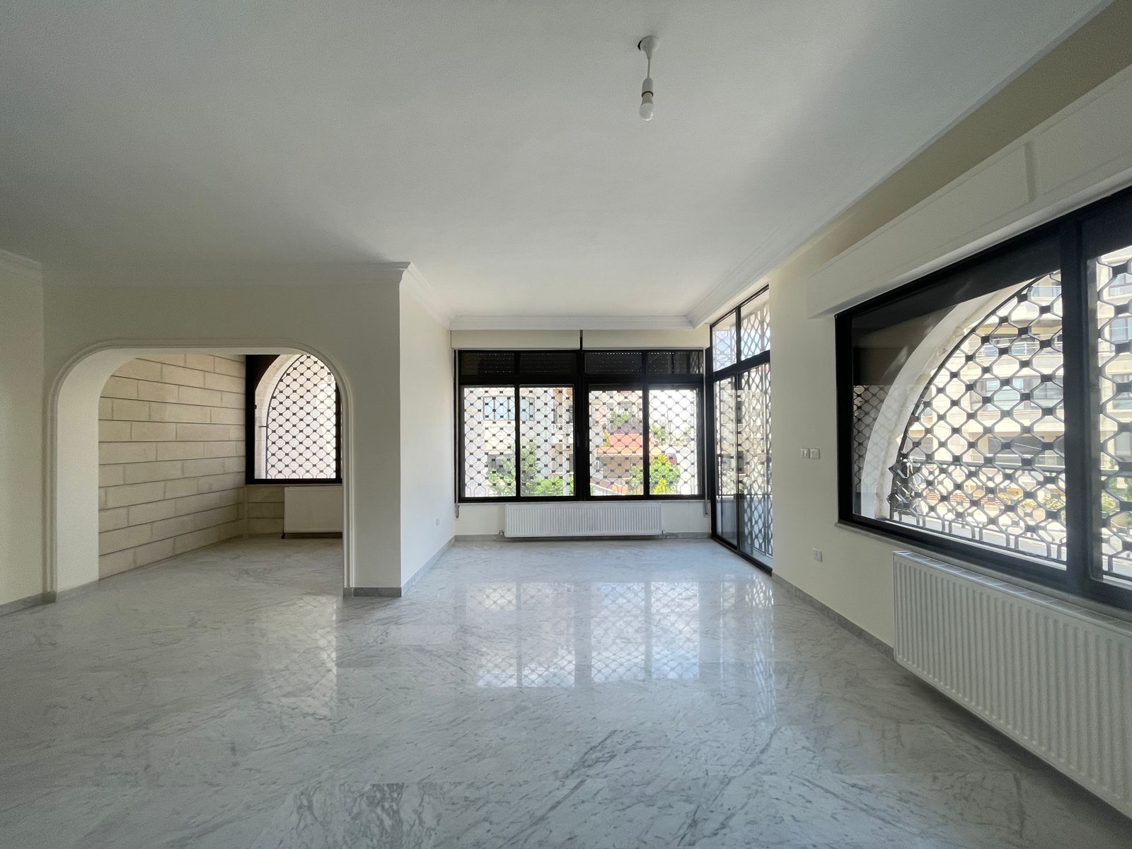 649567a7-691a-4c6a-b60a-18728edfcdb3 Empty bright living room with large windows, marble floor, and decorative metal grilles; archway to a side room.