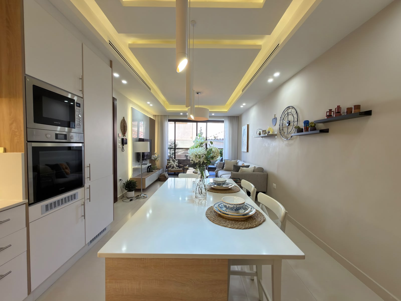 649863dd-7deb-4de0-b3b8-15315ad070be Open-concept kitchen and living area with white cabinets, built-in oven and microwave, a white island, and a dining table set with dishes and flowers.
