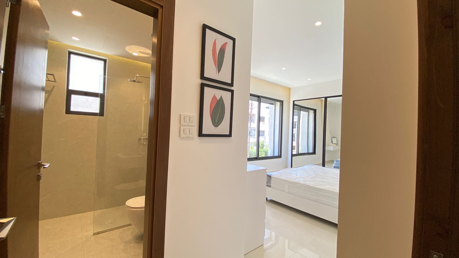 657526c0-d03f-448c-baad-d6ee134adbd7 Modern bedroom with white furniture, large windows, and two framed leaf art prints on a white wall; bathroom with glass shower visible to the left.