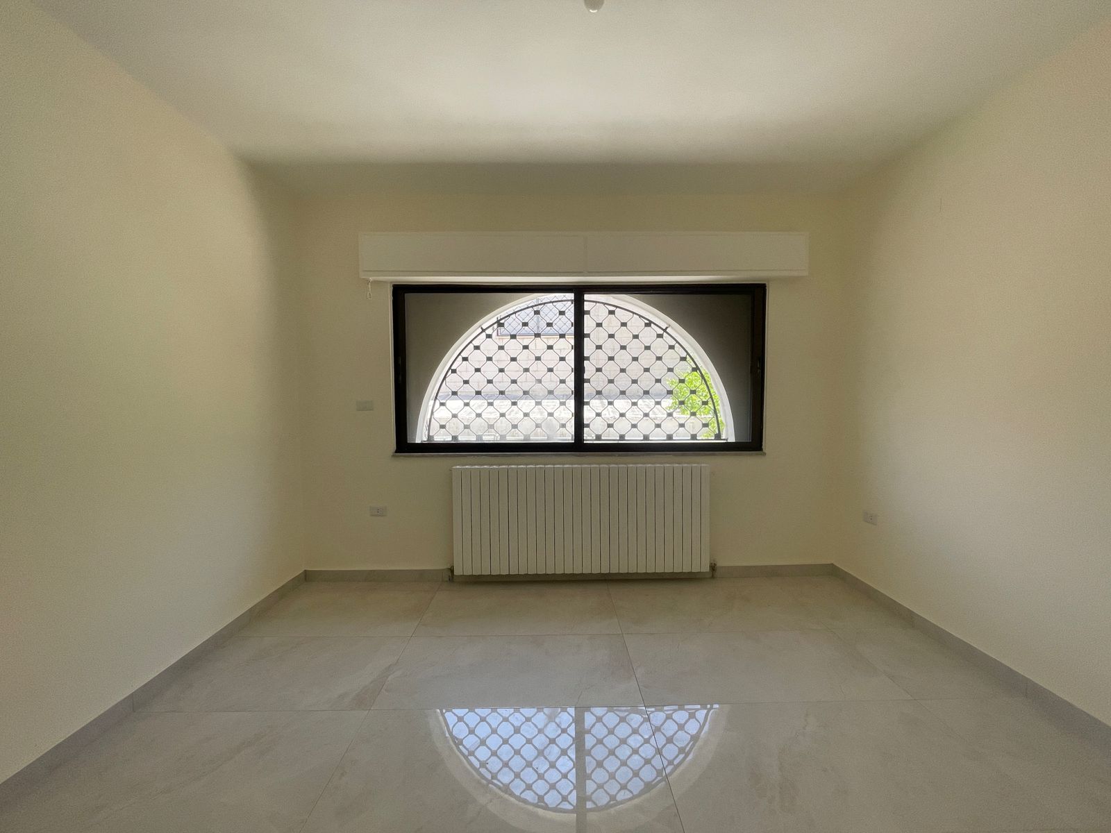 6580d9d6-ac40-4e95-9258-34f9d3bd6bc6 Empty rectangular room with a tall arched window featuring a metal lattice, radiator below, and glossy tiled floor reflecting light.