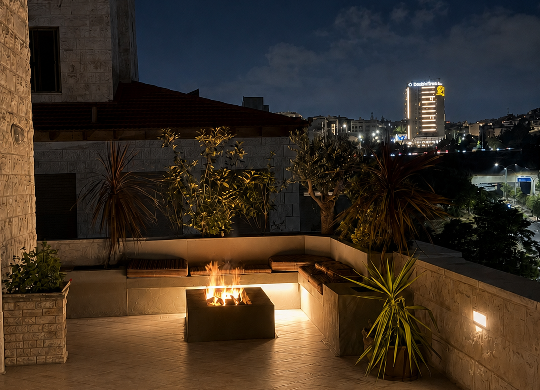Night view of a illuminated rooftop terrace with a square fire pit and built-in seating, potted plants, and stone walls against a dark city skyline.