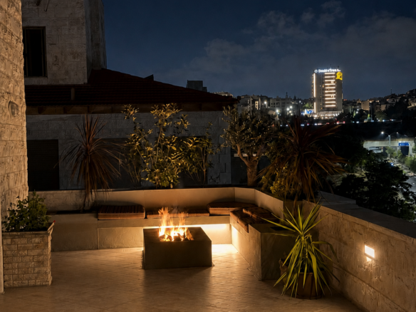 Night view of a illuminated rooftop terrace with a square fire pit and built-in seating, potted plants, and stone walls against a dark city skyline.