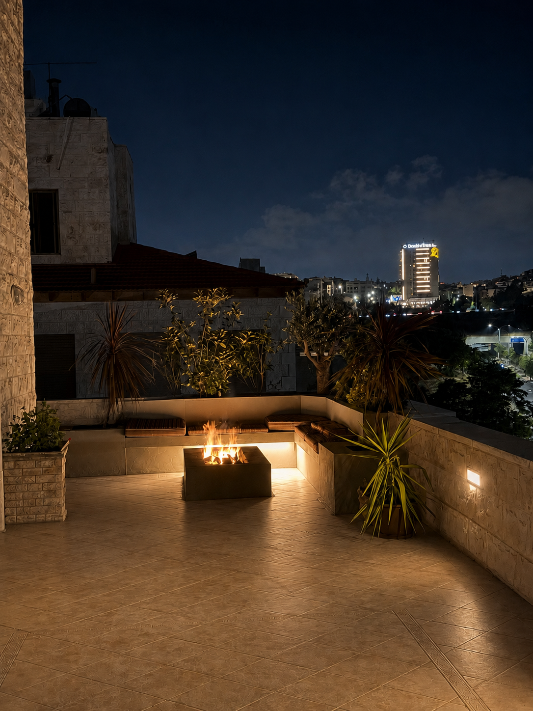 660583e2-56ad-44ae-812f-e9c105741c30 Night view of a illuminated rooftop terrace with a square fire pit and built-in seating, potted plants, and stone walls against a dark city skyline.
