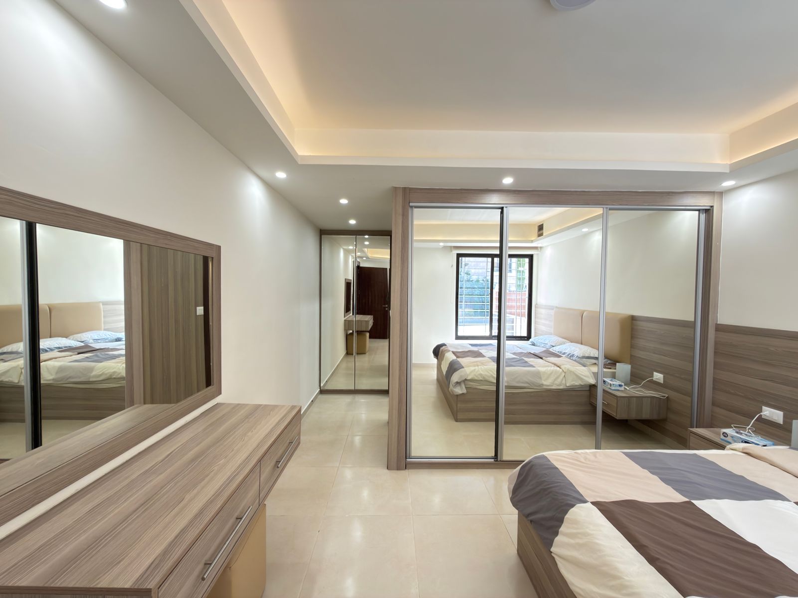 661a5b58-d60d-4ffd-b667-fbbc1b35583b Bright modern hotel bedroom with a large mirrored sliding-wardrobe, a wooden dresser, and a bed dressed in brown, beige, and white bedding; neutral tones and soft ceiling lighting create a calm space.