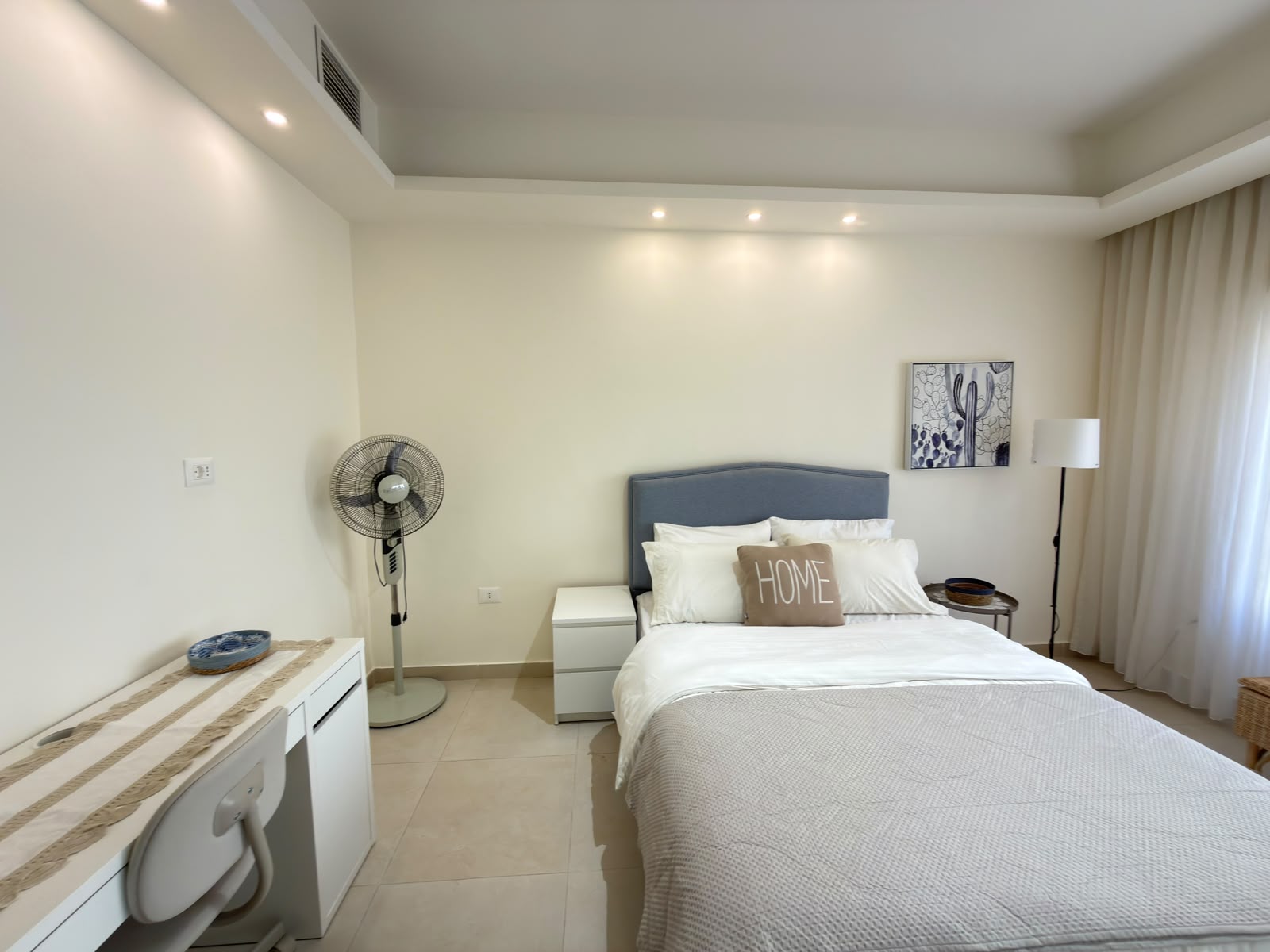 67d729b5-d740-4365-97c7-ea9aa7b3ecec Bright minimalist bedroom with a neatly made queen bed, gray headboard, white linens, and a decorative 'HOME' pillow by the window.