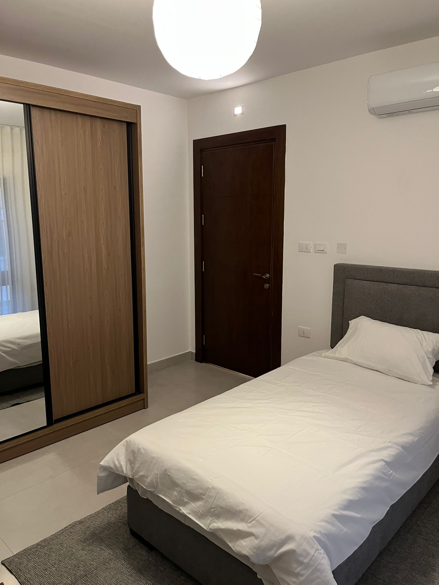 69d57961-41e3-4f47-af65-31f3e3ae97e2 Twin bedroom with two single beds in white linens, gray headboards, a wooden wardrobe with sliding mirrors, and a dark brown door; ceiling light overhead and an AC unit on the wall.