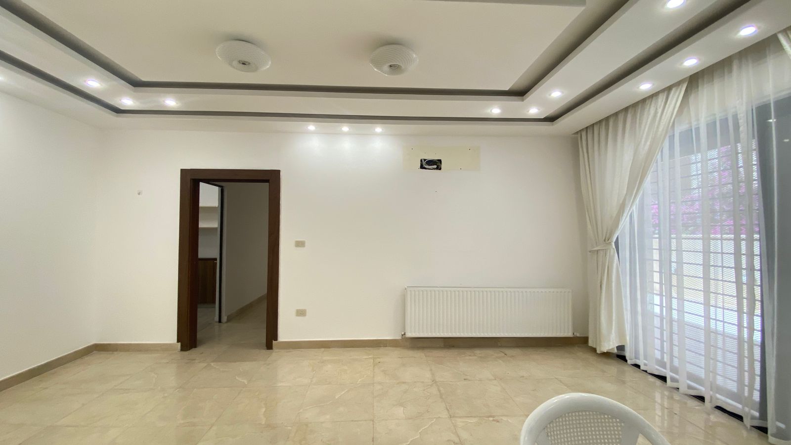 6b04ab00-165e-4a78-9ced-615114c8fdaf Empty living room with white walls and tiled floor, ceiling recessed lights, and a large window with sheer white curtains.