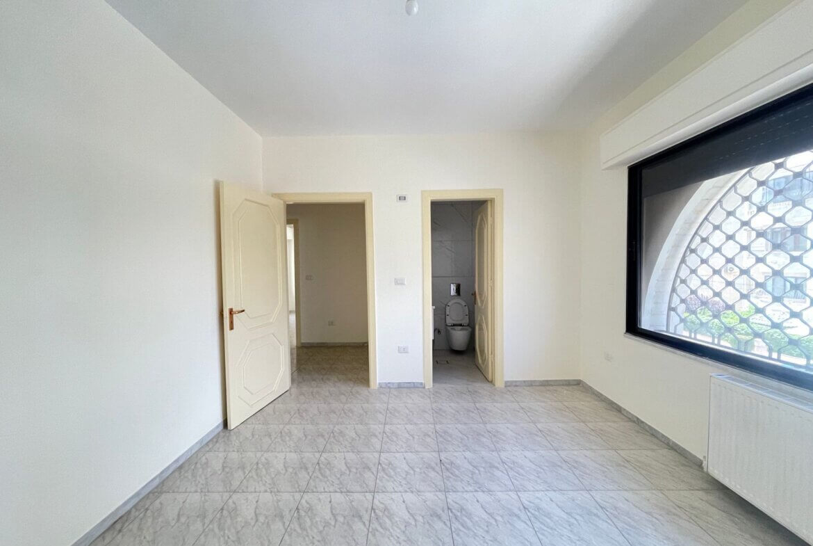 6b6900ea-787e-4505-a762-f4b595d37775 Empty light-filled room with tiled floor, two doorways (one to a bathroom) and a large window with a security grid on the right side, bare lightbulb hangs from the ceiling