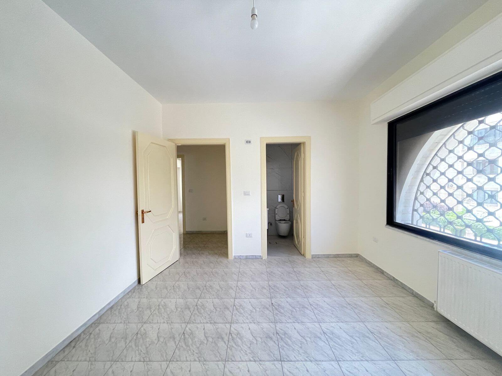 6b6900ea-787e-4505-a762-f4b595d37775 Empty light-filled room with tiled floor, two doorways (one to a bathroom) and a large window with a security grid on the right side, bare lightbulb hangs from the ceiling