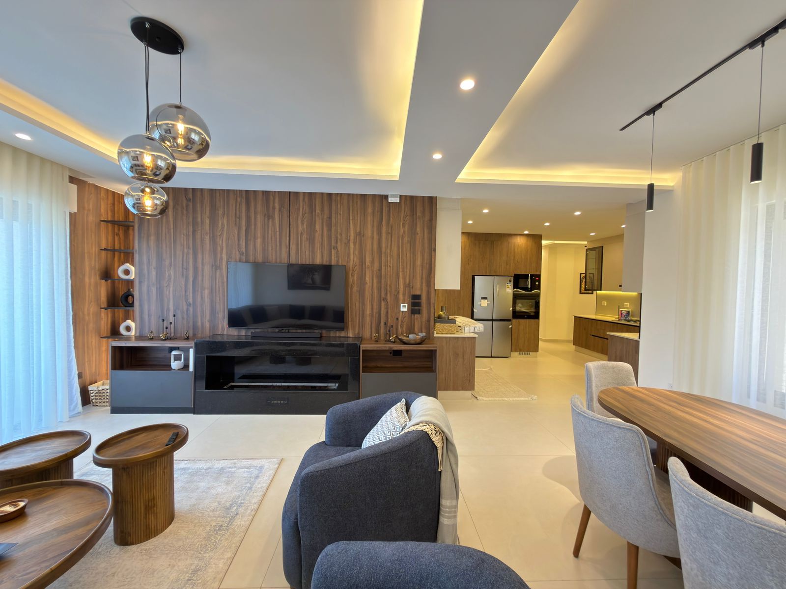 6e021781-6eb4-435e-bc2c-4deb2d918b75 Open-concept living room with a wood-paneled accent wall, mounted TV, and black fireplace cabinet; kitchen in the background and round wood coffee tables in the foreground.
