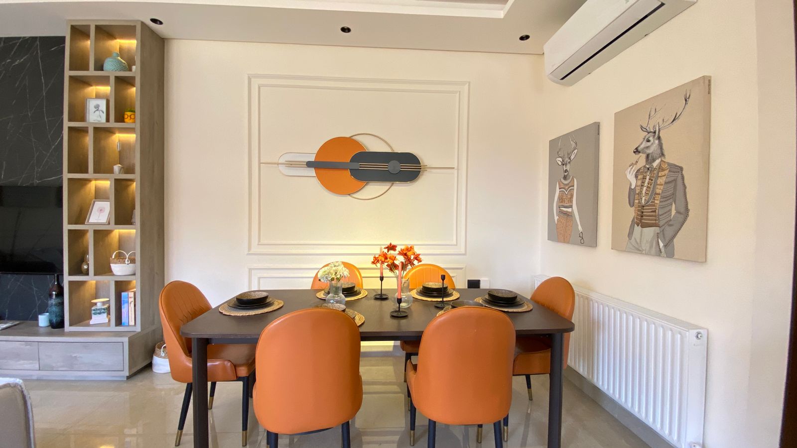 6e0cb120-1d5a-47a3-8ba2-e8f09e9d084a Dining area with six orange chairs around a dark rectangular table, set with plates and flowers, against a modern wall sculpture.