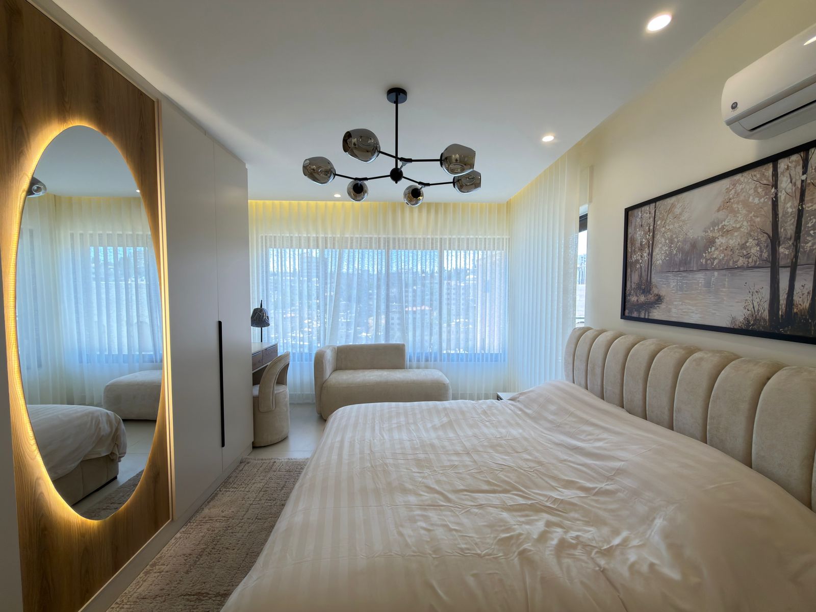 6e5c1f14-e4e8-460a-b377-521b8ed3af4e Modern bedroom with a long beige upholstered headboard, a white bed, and a large circular-molded mirror with warm backlighting on the left.