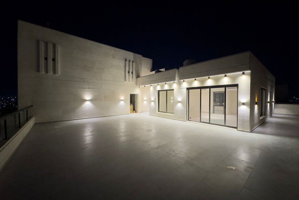 Nighttime rooftop terrace of a modern building with warm wall lights and large sliding glass doors, beige stone walls. An open doorway and city lights in the distance.